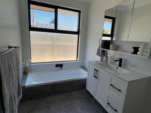Same bathroom after renovation: white vanity, mirror cabinet and tiled bath