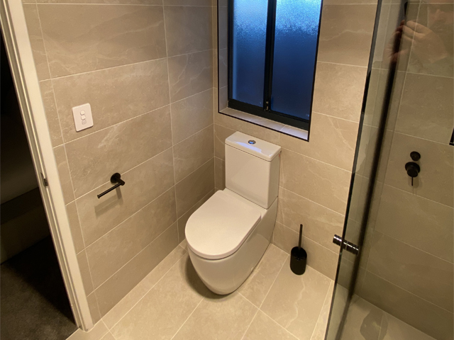 Same bathroom after renovation: grey wall and floor tiles, toilet and black fittings