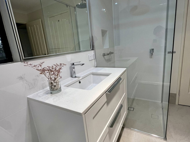 White vanity with stone top below a wall mirror, beside a frameless glass shower
