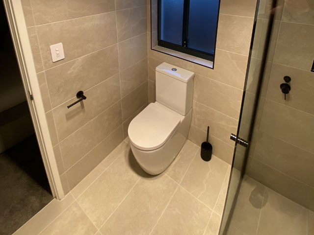 Same bathroom after renovation: grey wall and floor tiles, toilet and black fittings